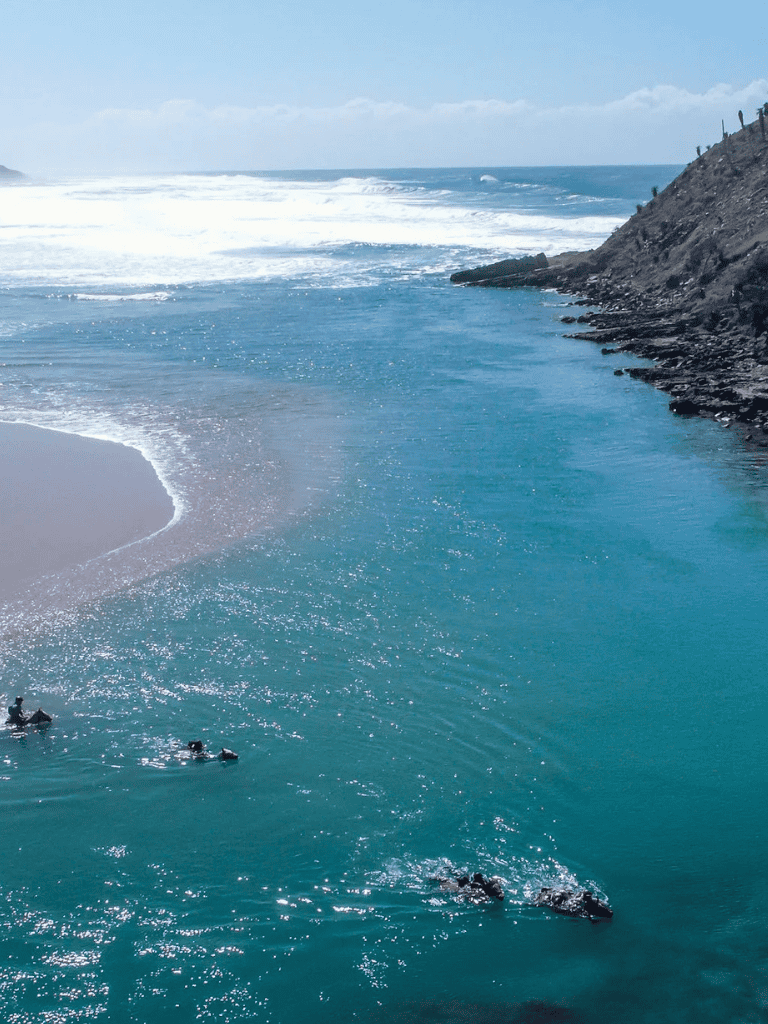 Bright blue ocean water with surfers and rocky coast at a scenic seaside location.