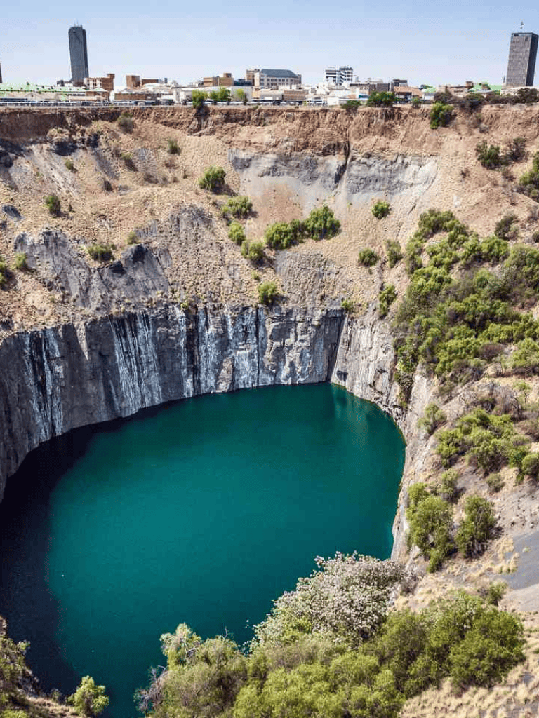 Vast quarry with a turquoise lake surrounded by rocky cliffs and sparse greenery, city skyline in background.