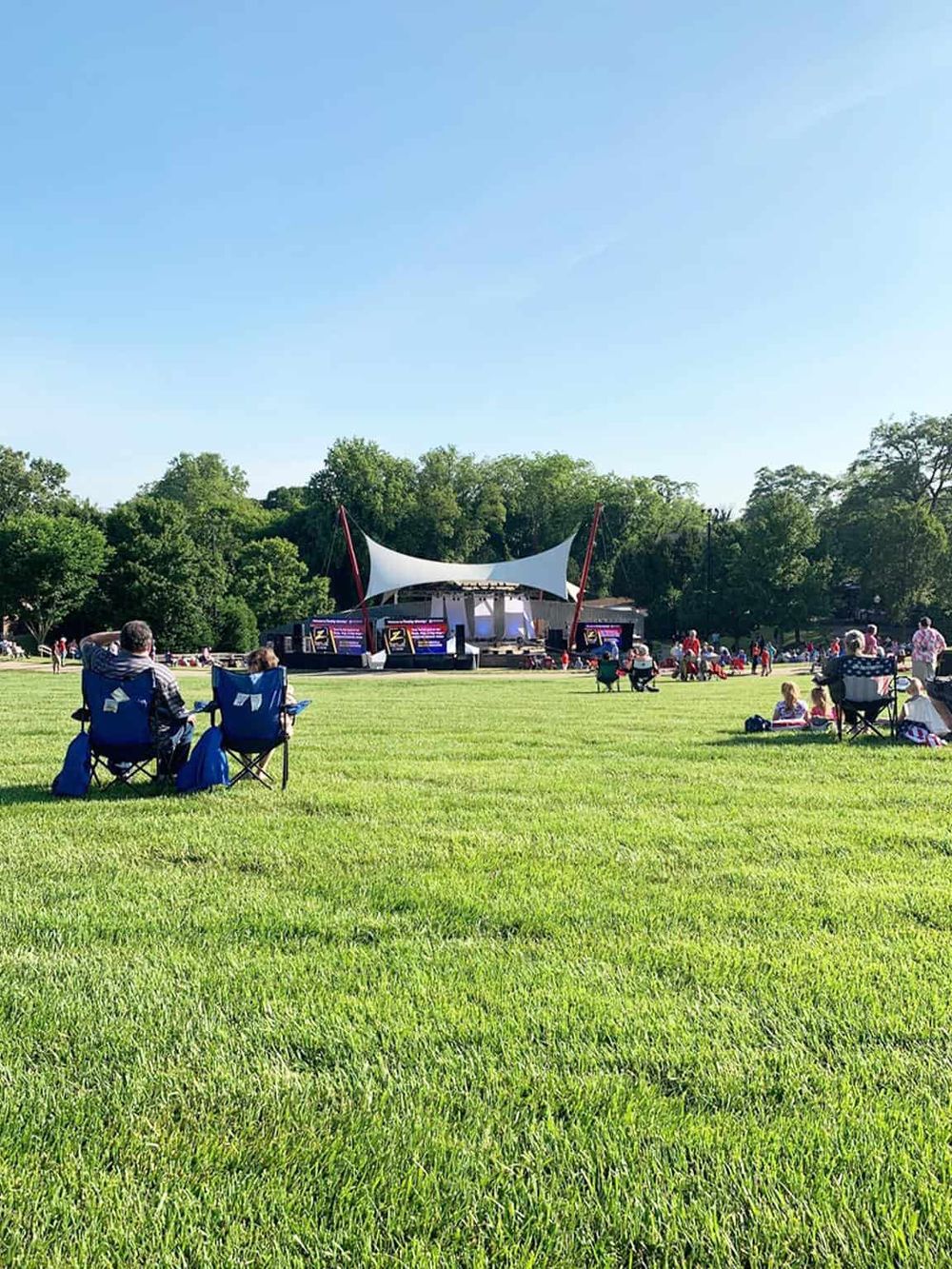 Outdoor concert in a park with people sitting on chairs, enjoying music under a bright blue sky.