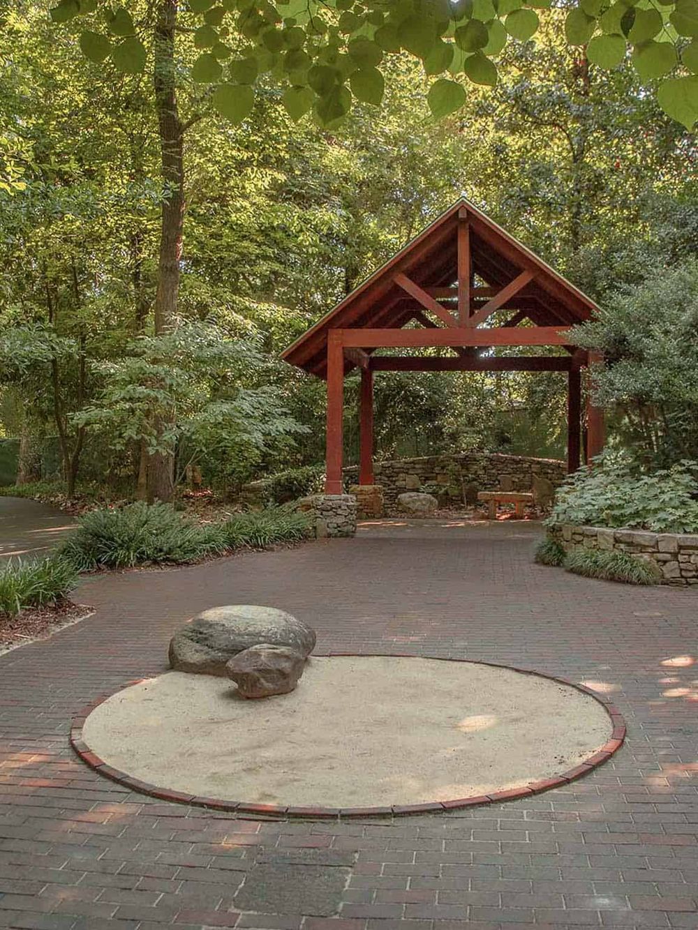 Serene garden pavilion with red wooden structure amidst lush green trees and a brick pathway.