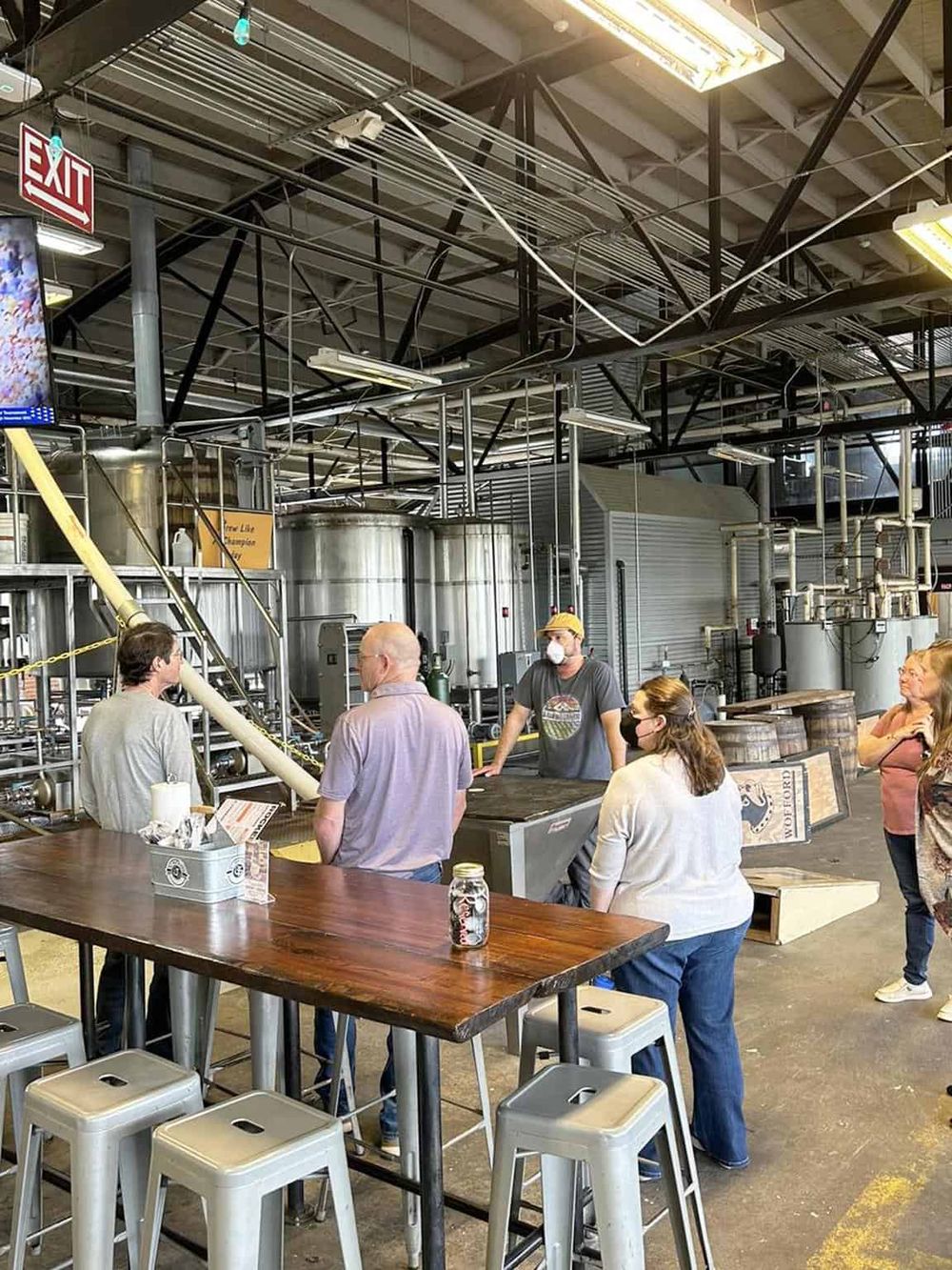 A brewery tour with visitors and a guide at a craft brewery, industrial tanks in the background, and people wearing masks.