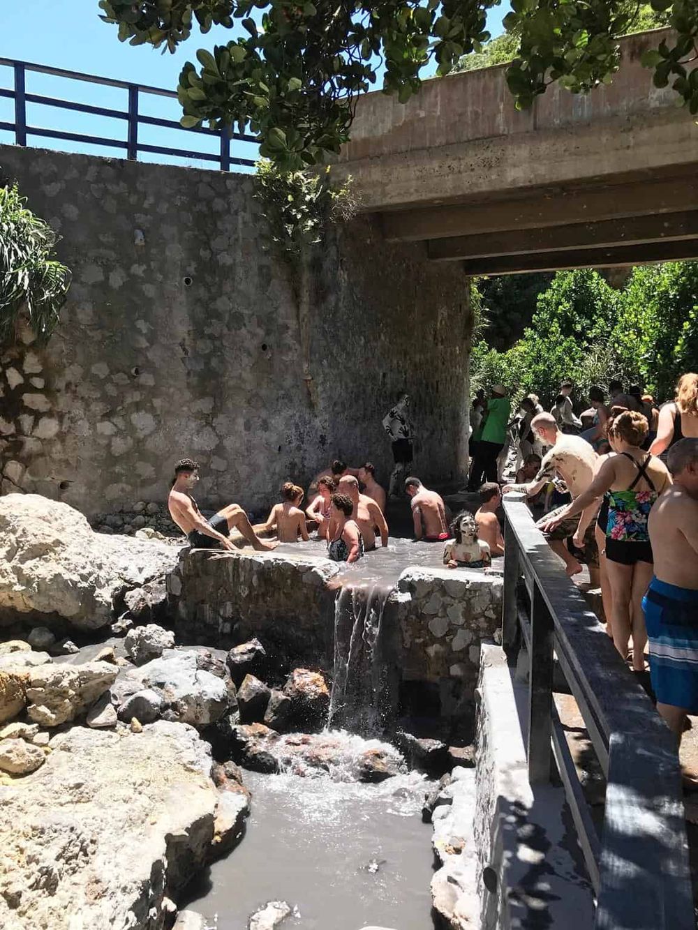 People enjoying hot springs under a bridge in a scenic outdoor setting.