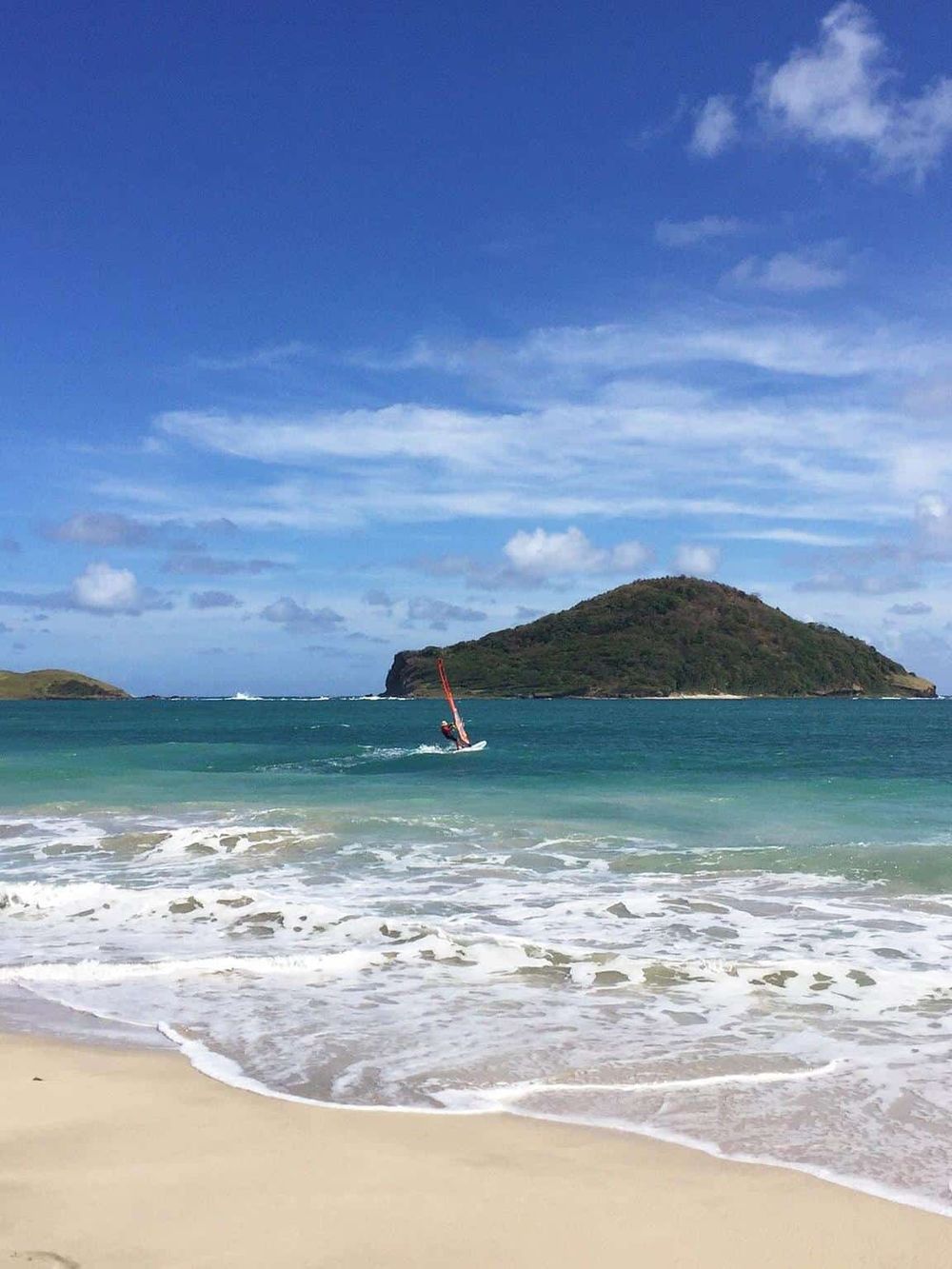 Stunning beach scene with a windsurfer on turquoise waters and lush island in the background.