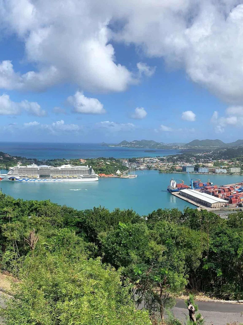Stunning view of the turquoise harbor with cruise ships, cargo containers, and lush green hills in the background.