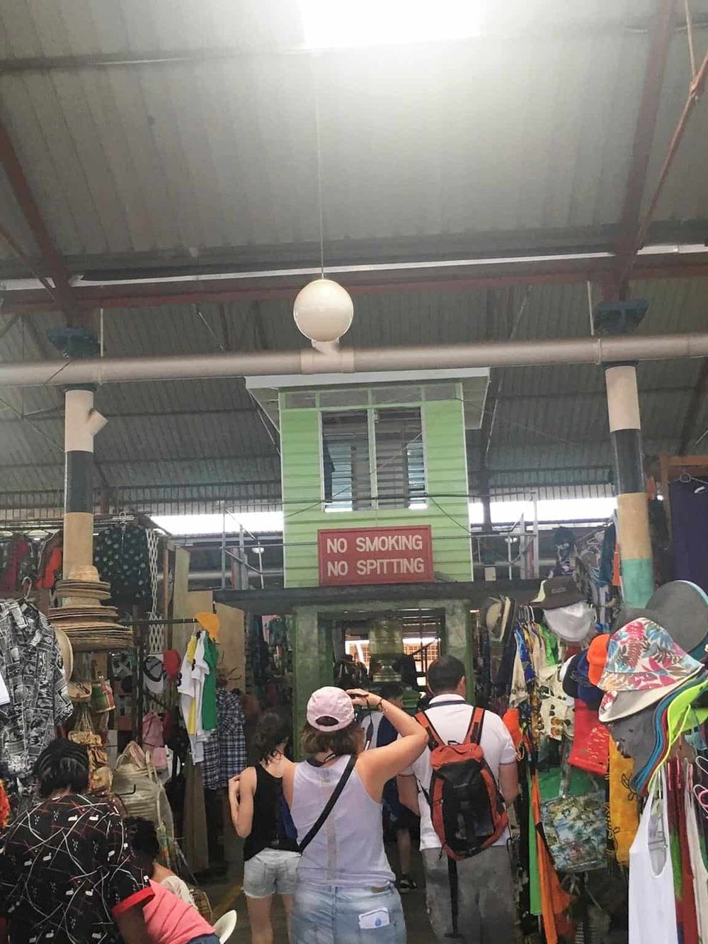 Busy indoor market with diverse shoppers and clothing stalls, vibrant atmosphere, and a "No Smoking No Spitting" sign.