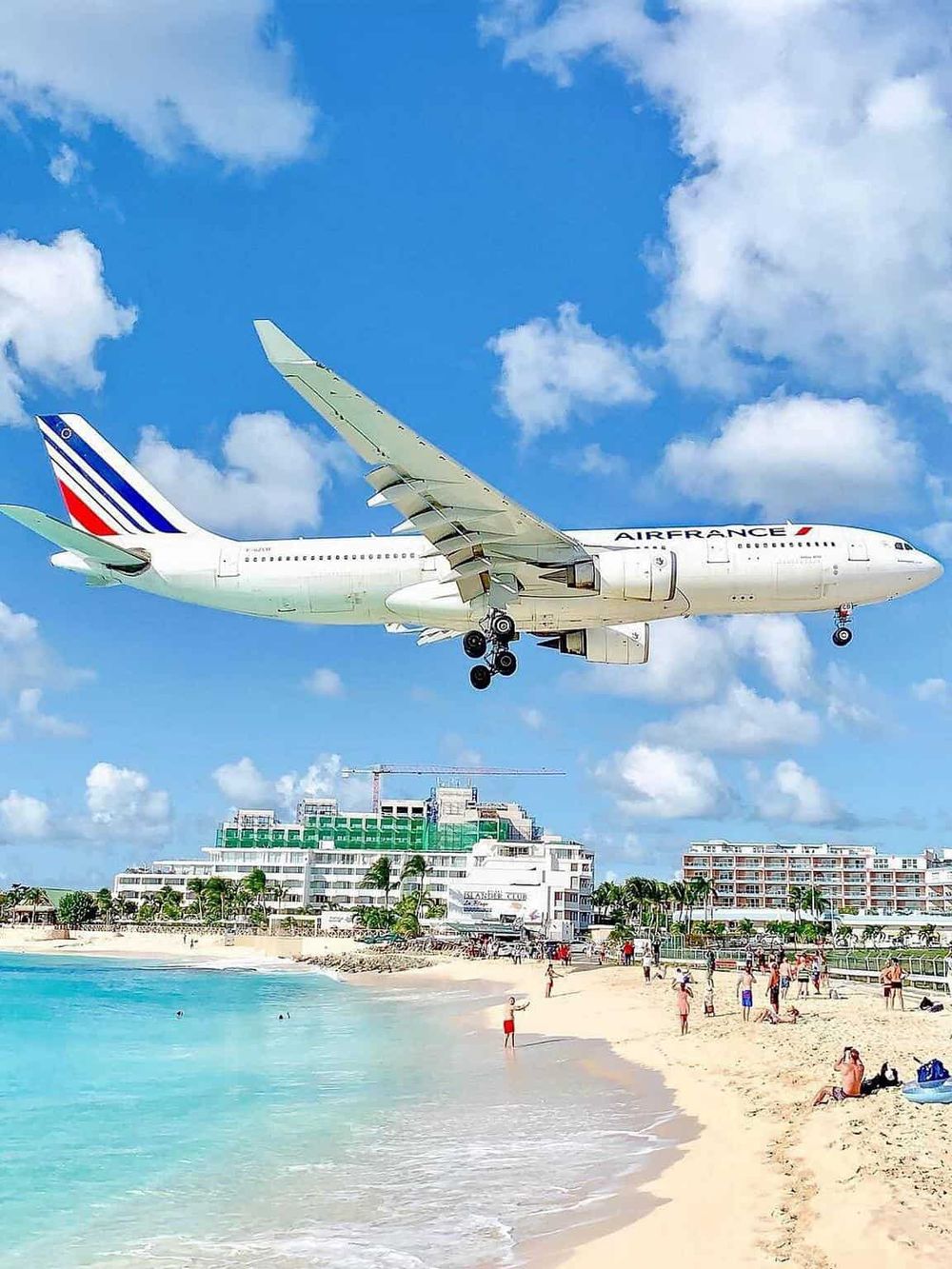 Modern Air France airplane landing above a tropical beach resort, white sand, clear blue water, and vacation travelers.