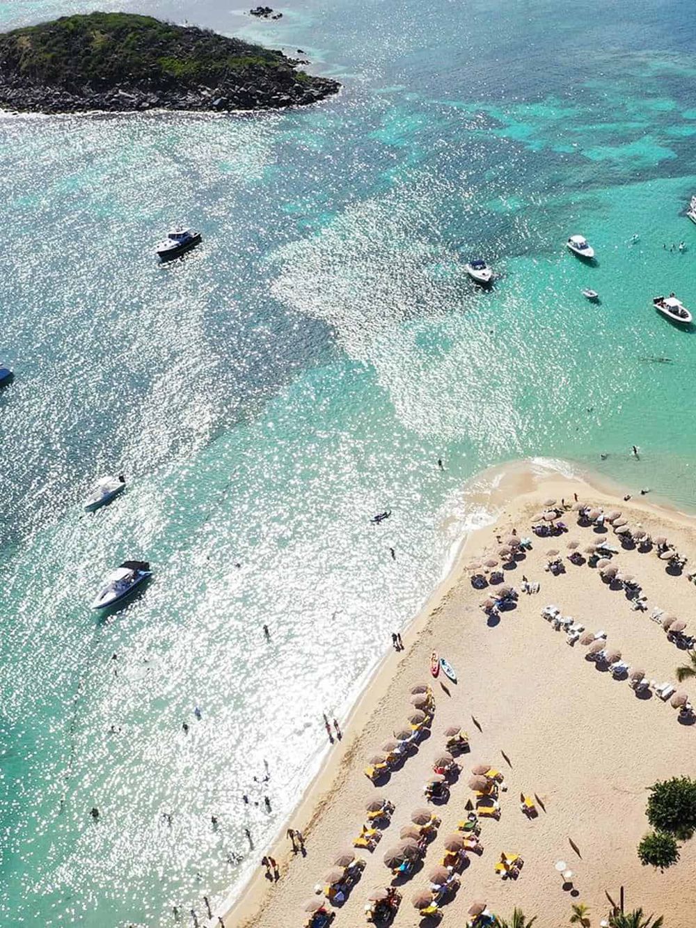 Aerial view of turquoise beach with boats, umbrellas, and sunbathers, showcasing leading travel destinations and water activities.