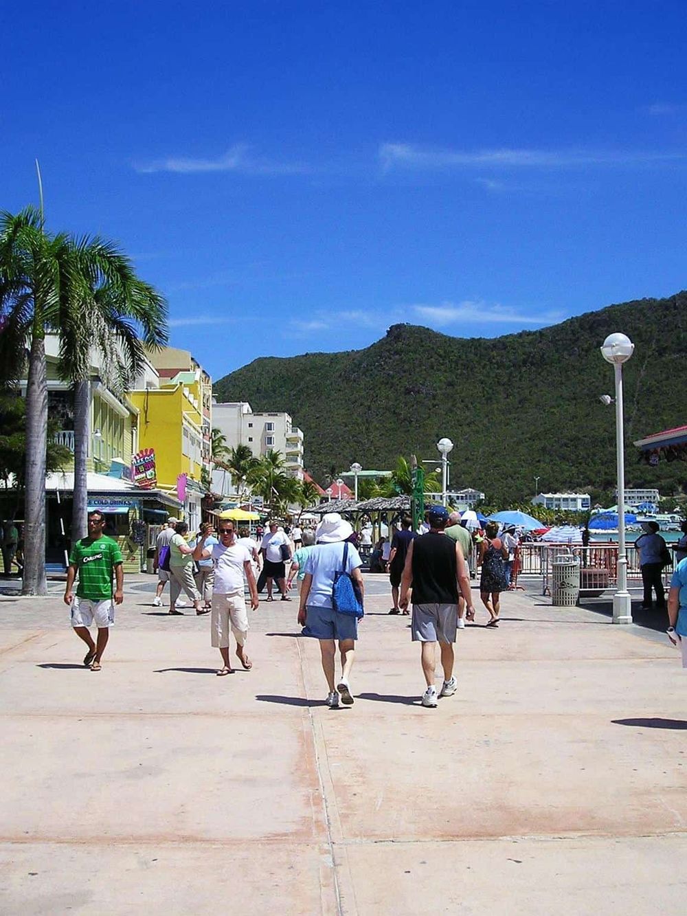 Vibrant seaside promenade with tourists, colorful buildings, palm trees, and scenic mountain views.