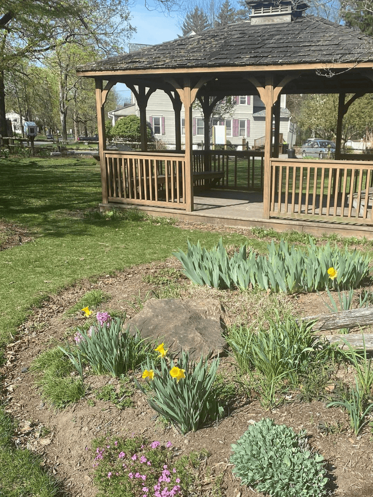Charming wooden gazebo in a lush garden with blooming flowers and community homes in the background.