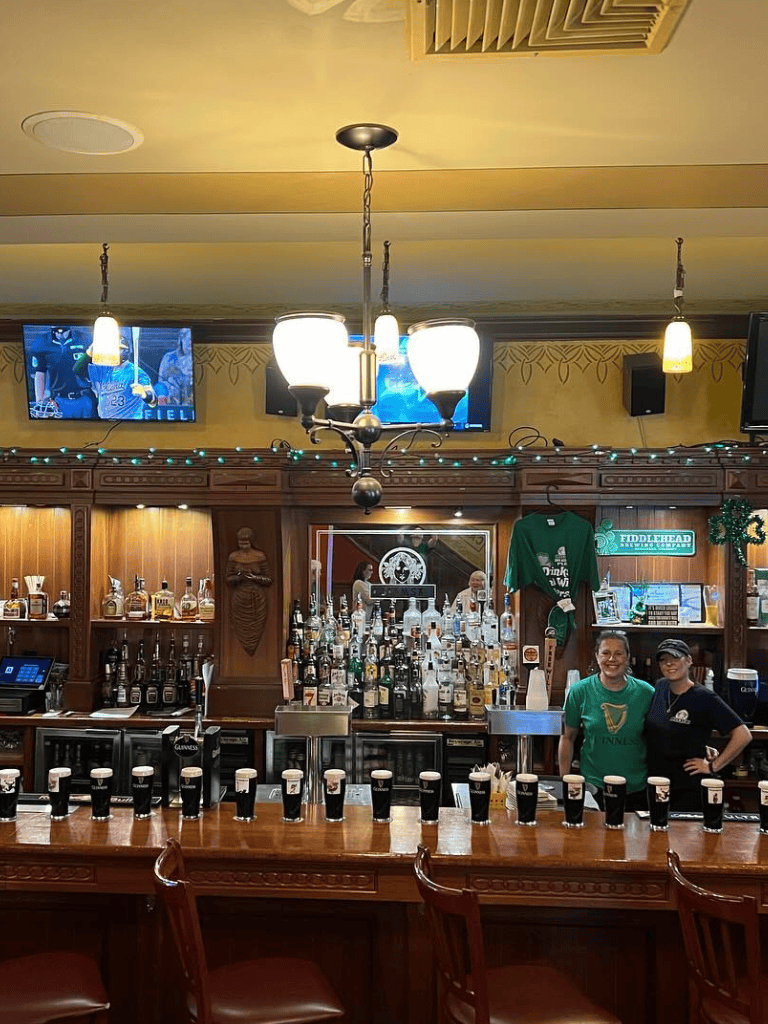 Beer bar with pints, TVs showing sports, and staff in green shirts in a cozy pub setting.