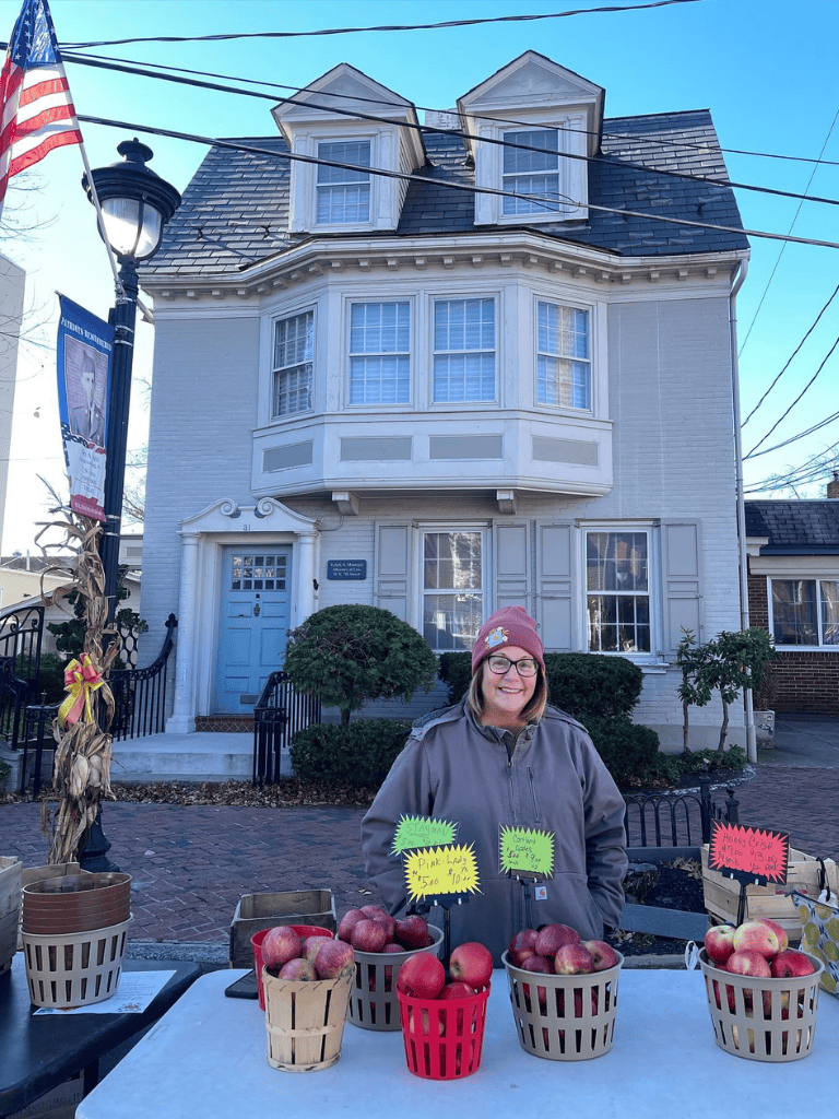Fresh apples for sale at a local market stand, with a charming historic house in the background. Perfect for supporting local farmers and community events.