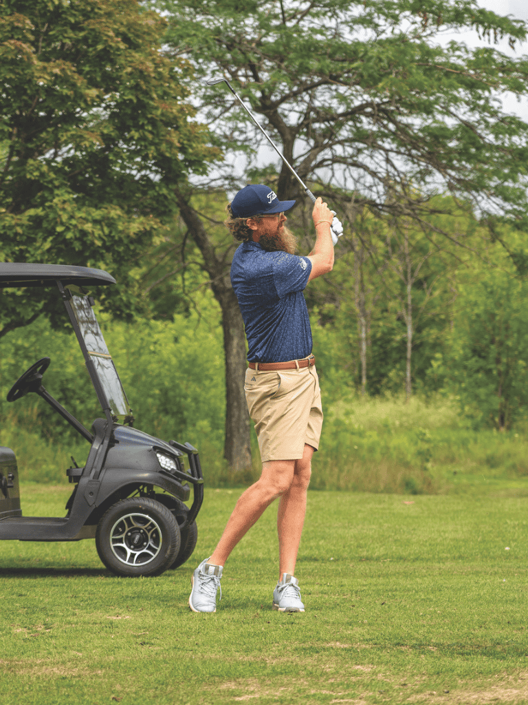 Golf player swinging club on lush golf course with golf cart in background.