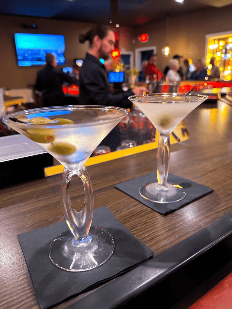 Olive martini cocktails served at a bar with bartenders and patrons in the background.
