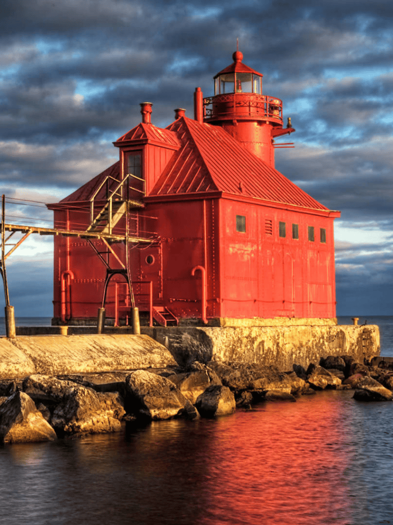 Lighthouse along rocky shoreline at sunset with dramatic sky, scenic coastal navigation point.