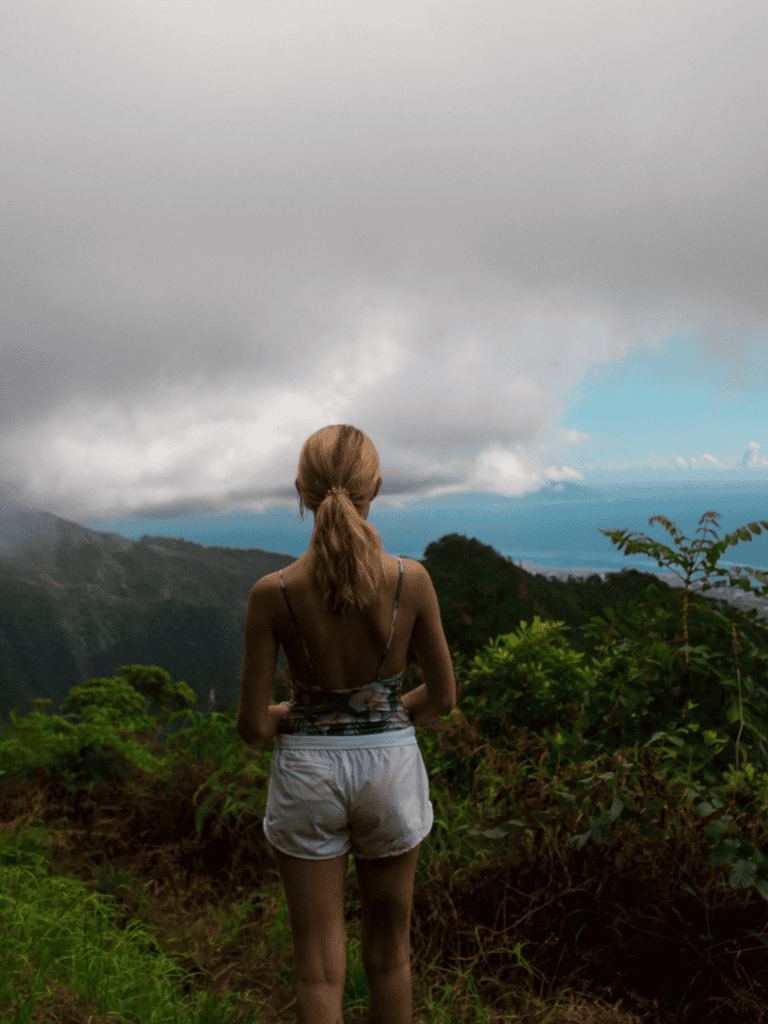 Girl standing on mountain trail overlooking lush green mountains under cloudy sky.