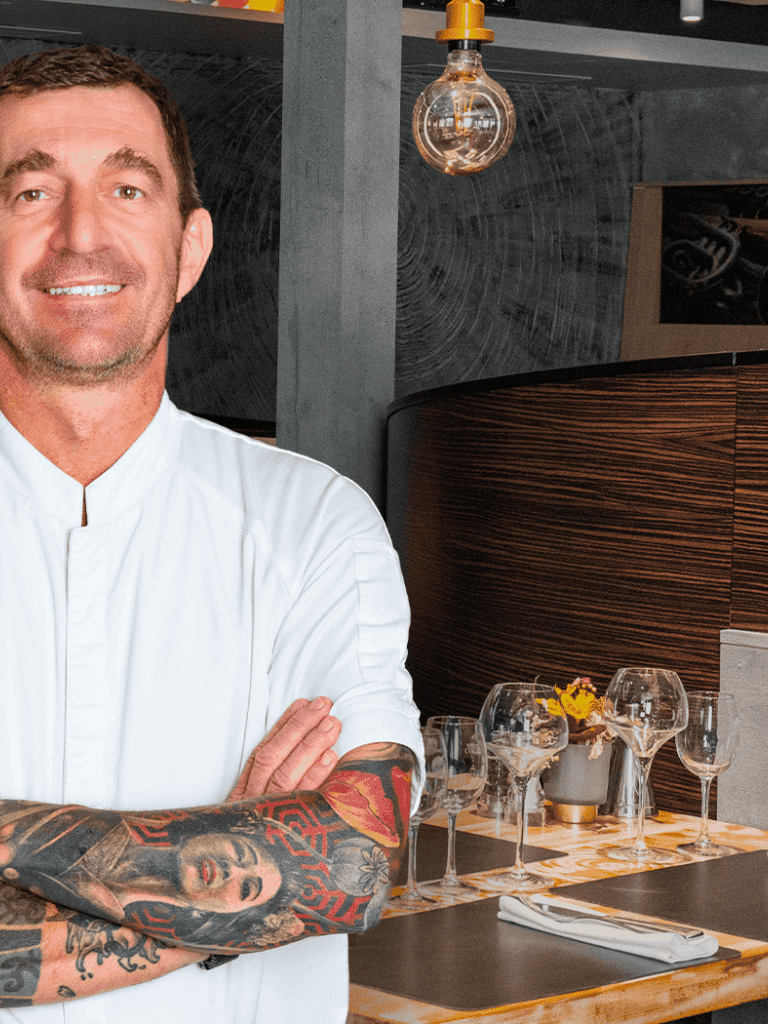 Chef in professional kitchen with tattoos, modern dining setup, cozy ambiance, and stylish decor.