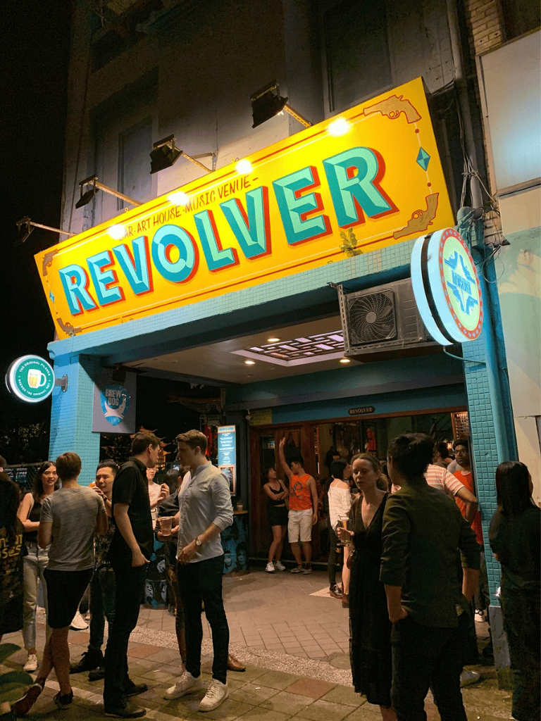 Vibrant bar entrance at Revolver venue with a lively crowd, neon signage, and outdoor nightlife scene.