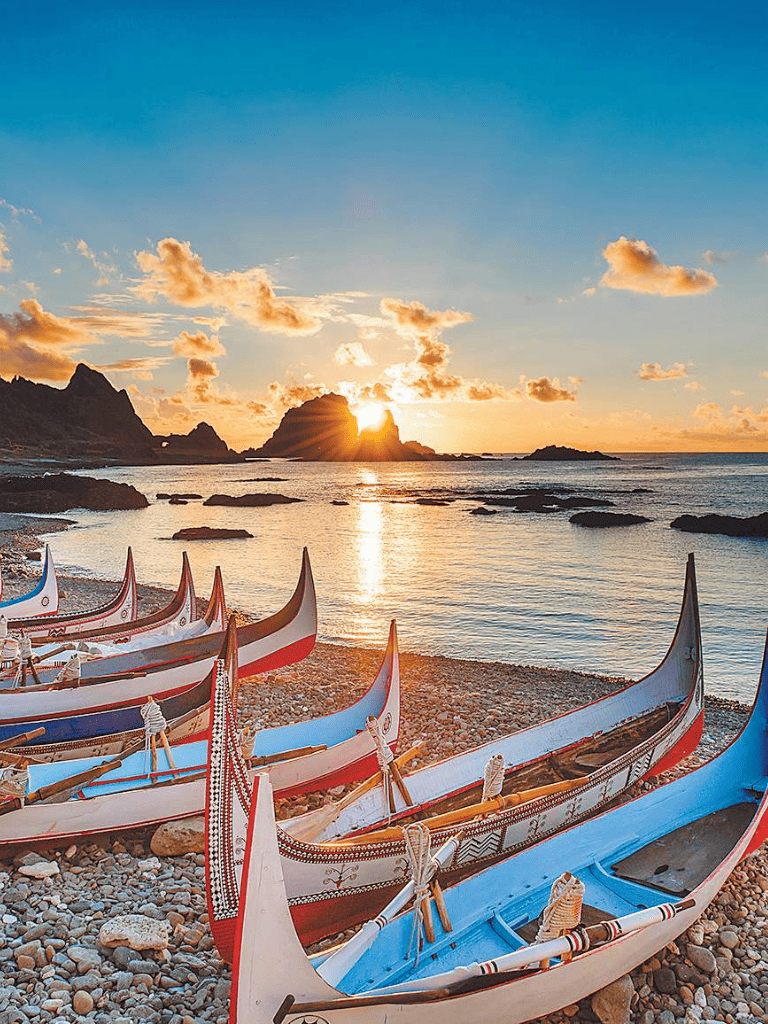 Colorful traditional boats on rocky beach at sunset with rocky islands in background, scenic coastal view.