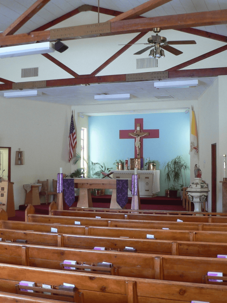 Quiet church interior with pews, altar, crucifix, flags, and ceiling fan for religious community gatherings.