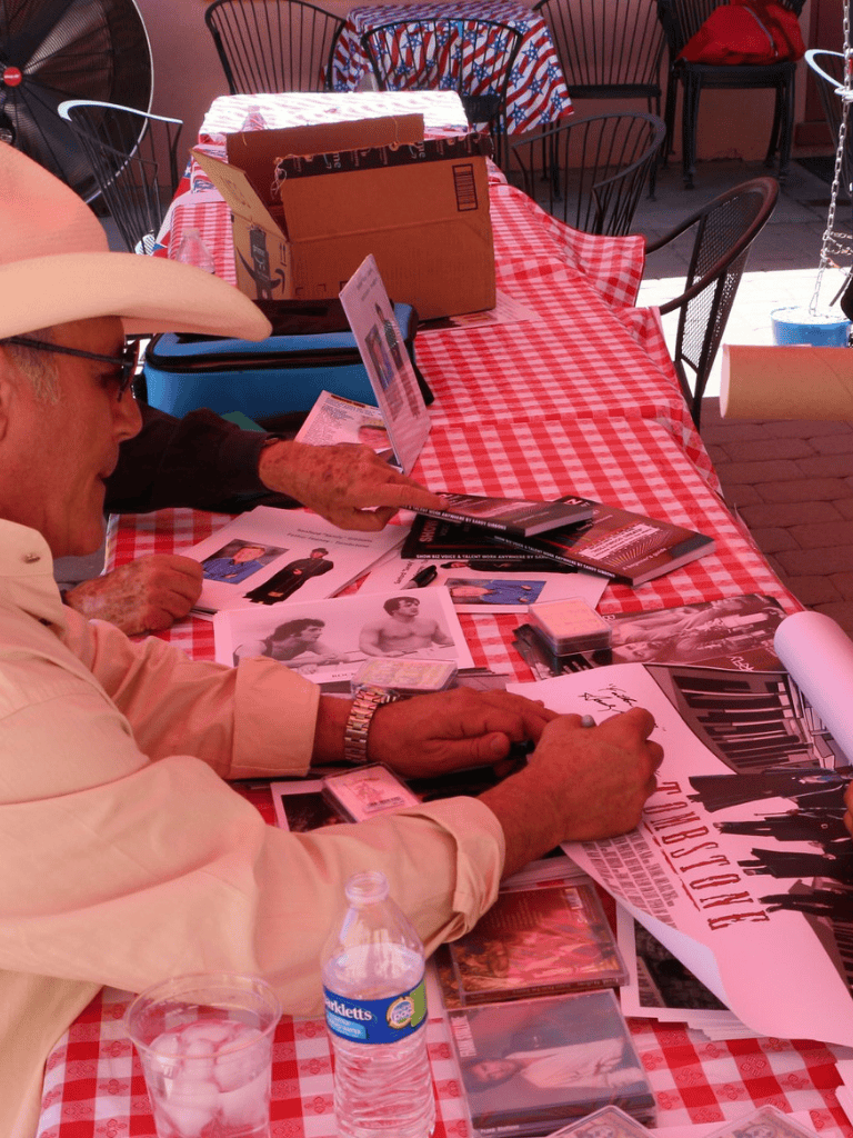 Vintage photo of a man wearing a cowboy hat signing posters at a table with red checkered tablecloth, memorabilia, and photos.
