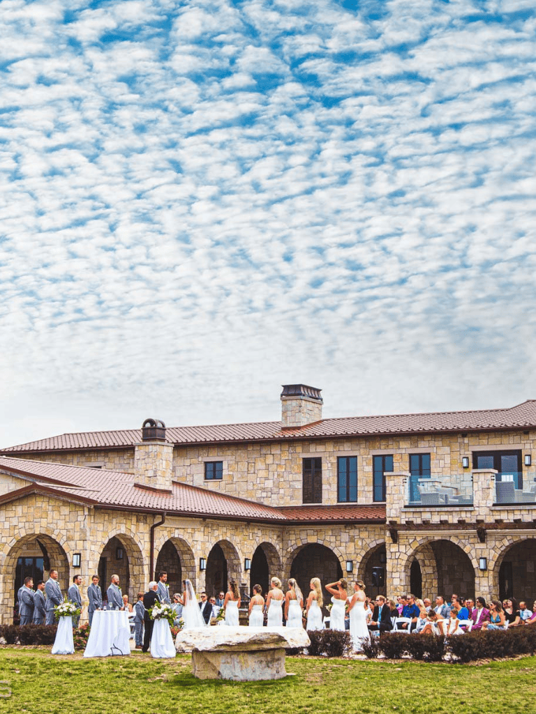 Elegant outdoor wedding ceremony under a scenic sky at a stone venue with guests and wedding party.