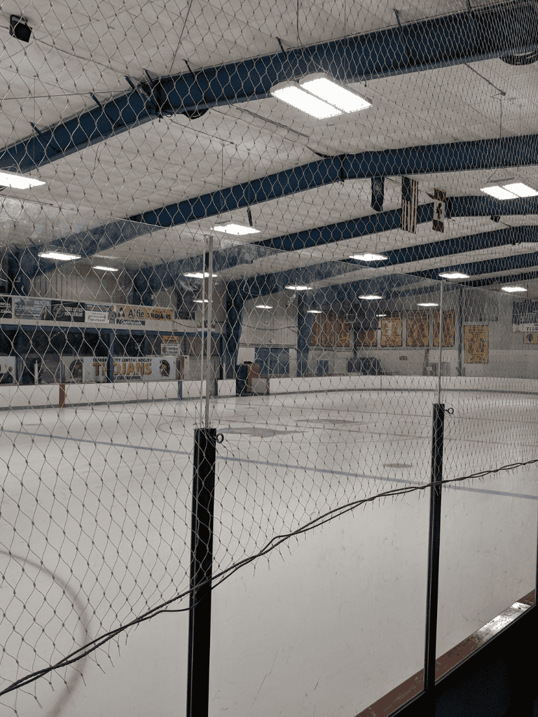 Shattered safety net at indoor hockey rink, empty ice skating arena with banners and overhead lights.