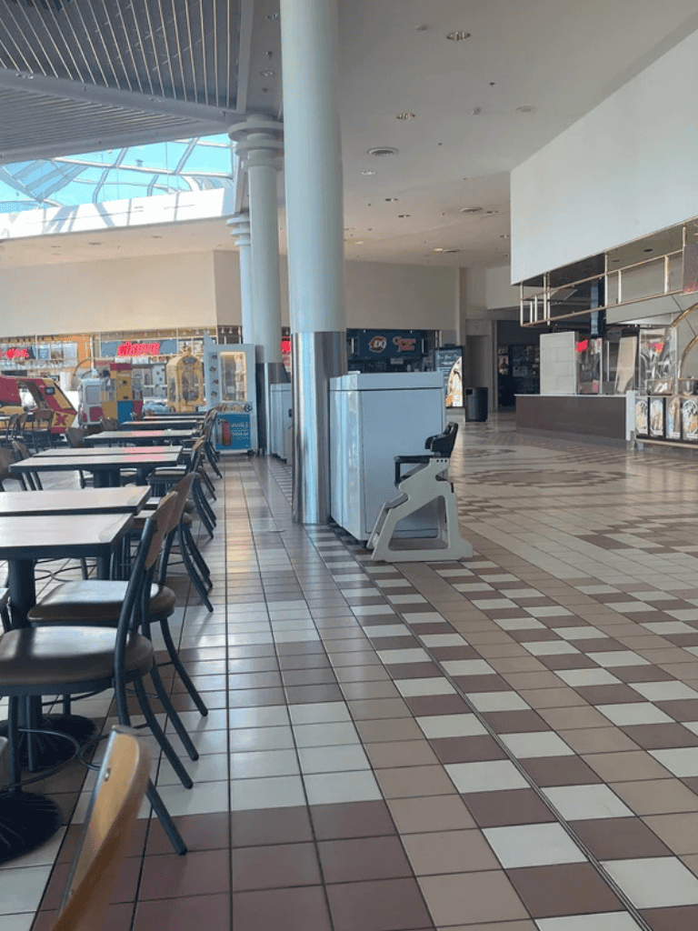 Empty shopping mall food court with tables and chairs, bright natural light, modern architecture, and retail stores in background.