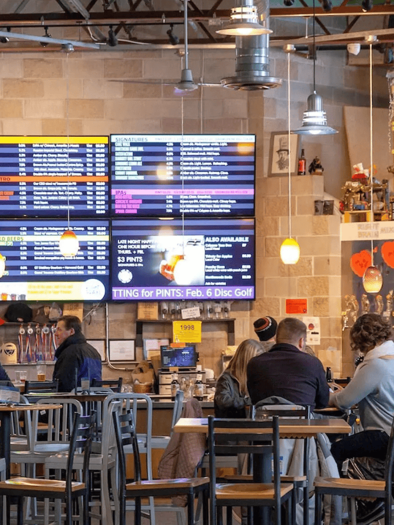 Digital menu boards at a lively bar or restaurant with patrons, featuring drink specials and event info.