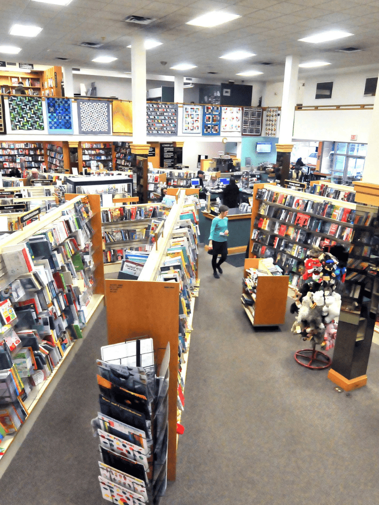 Bookshelf and customer in bookstore aisle with wall art decor, cozy shopping environment, wide selection, and comfortable browsing space.