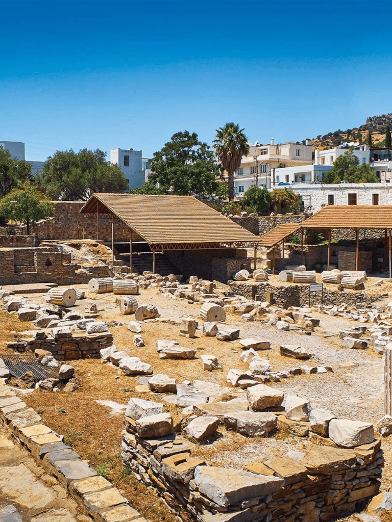 Ancient archaeological ruins with stone structures and modern buildings in the background, sunny day.