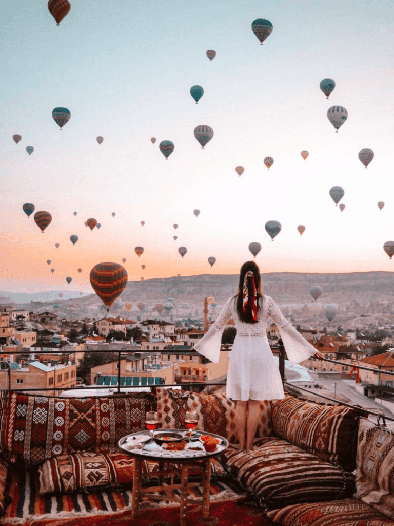 Hot air balloons over Cappadocia at sunrise, with a woman enjoying a scenic terrace view.
