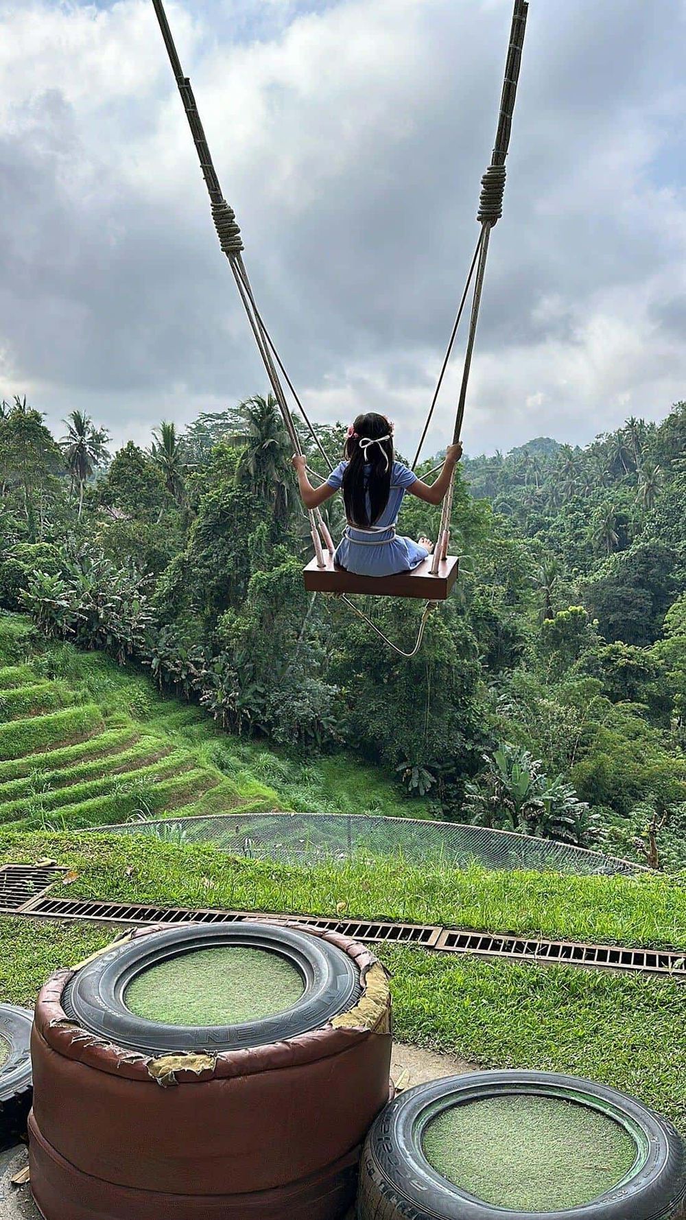 A girl swinging over lush green tropical landscape with terraced fields and dense trees.