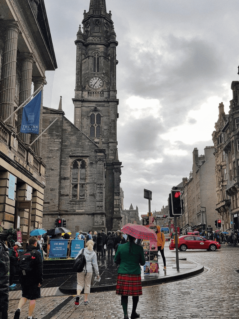 Colorful city street scene with historic clock tower, pedestrians with umbrellas, and rain-soaked cobblestones.