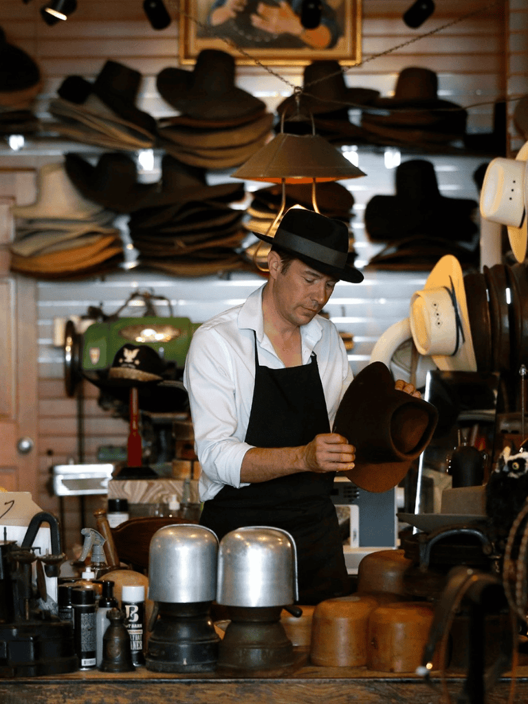 Hat shop with shelves of fedora and straw hats, vintage decor, and a man trying on a hat.