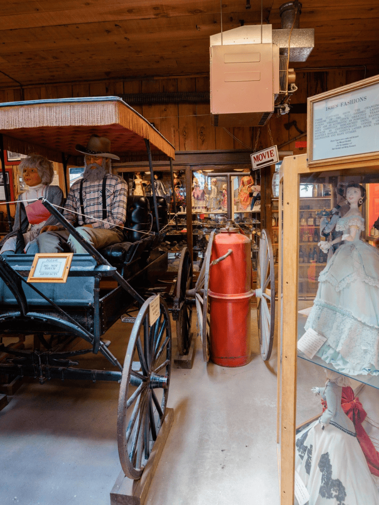 Antique horse-drawn carriage inside a vintage collectibles shop with doll mannequins and historical artifacts.