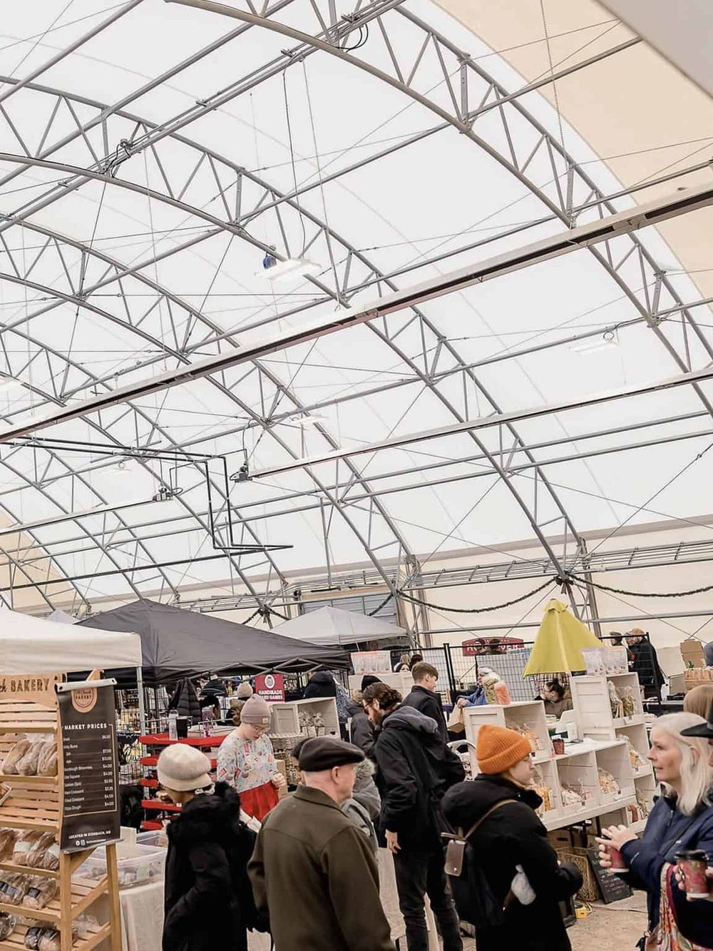 Vibrant indoor farmers market with diverse vendors and shoppers under a large, airy tent.
