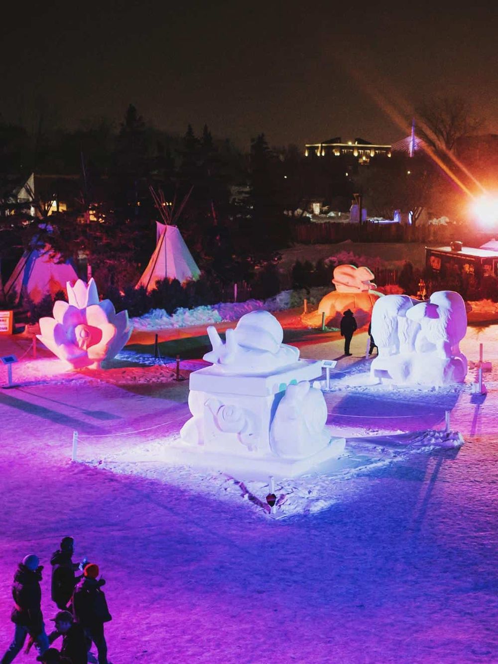 Colorful snow sculptures illuminated at night with illuminated tipi tents in the background.