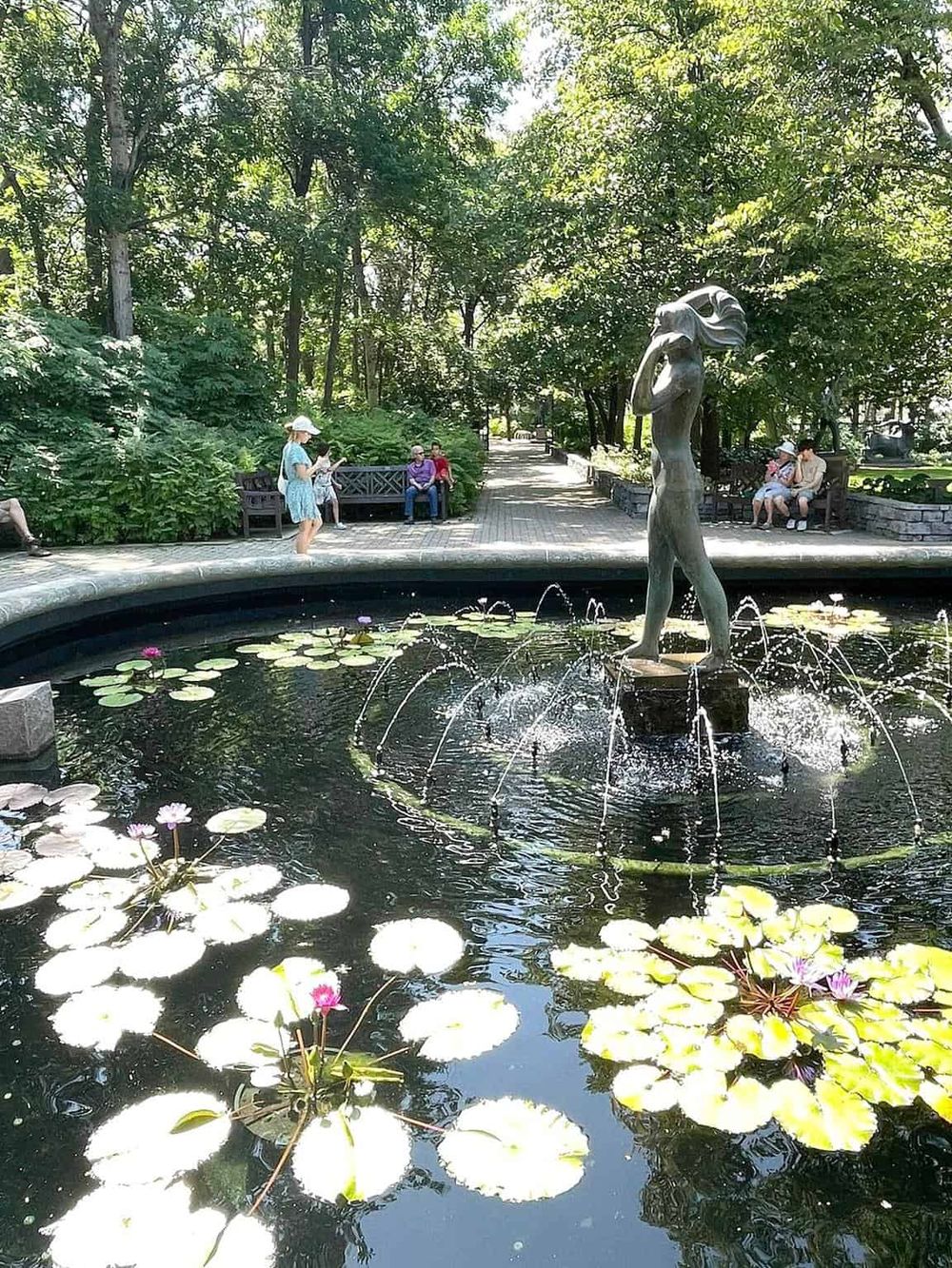 Serene park fountain with a female sculpture surrounded by lily pads and blooming flowers, ideal for relaxation.