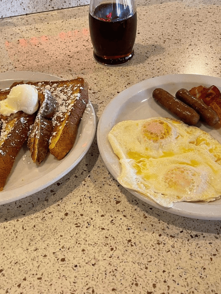Cream French toast with butter and powdered sugar, accompanied by fried eggs, sausage links, and bacon with a glass of soda.
