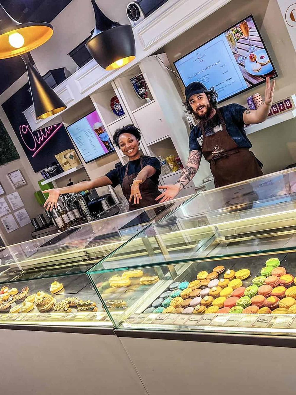 Brightly lit bakery display case with colorful macarons and pastries, cheerful staff presenting delicious desserts.