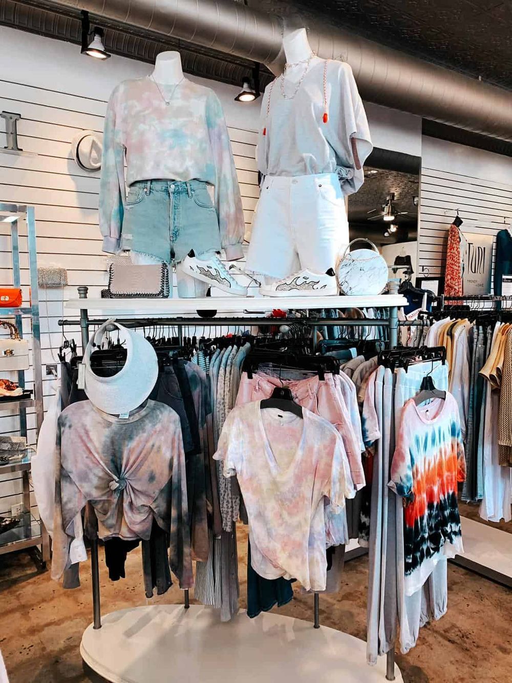 Colorful casual clothing store display featuring tie-dye shirts, denim shorts, and sneakers.