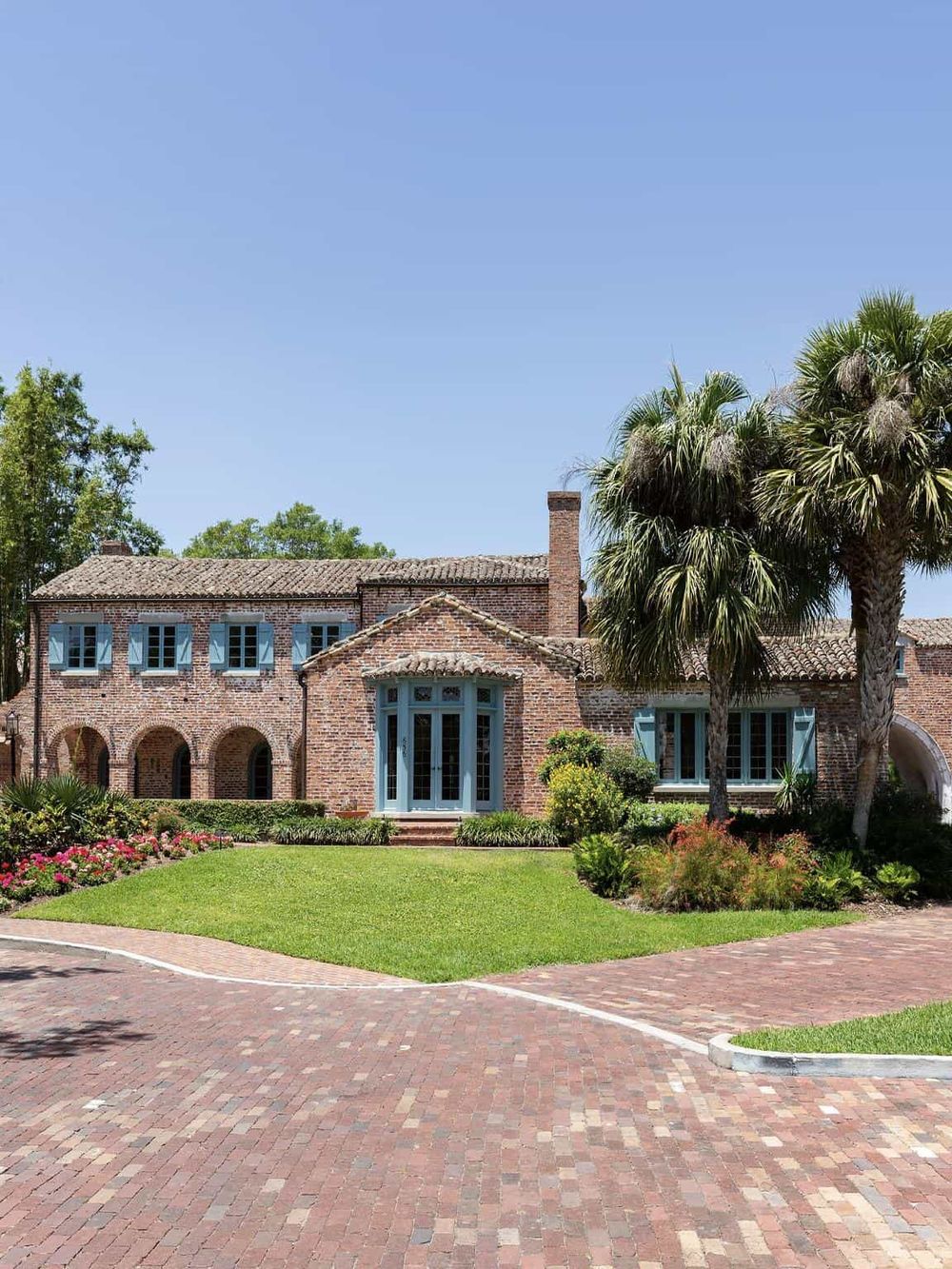 Charming brick house with blue accents, lush landscaping, palm trees, and a paved driveway, perfect for directions and travel.