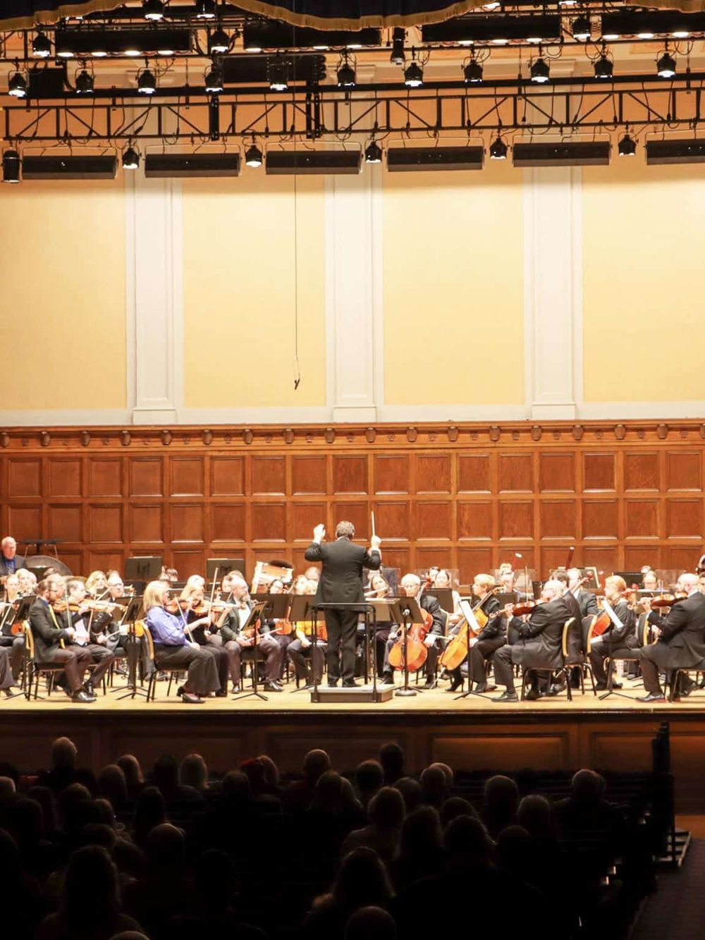 Orchestral concert in a large hall with musicians and conductor on stage, audience enjoying a live classical music performance.