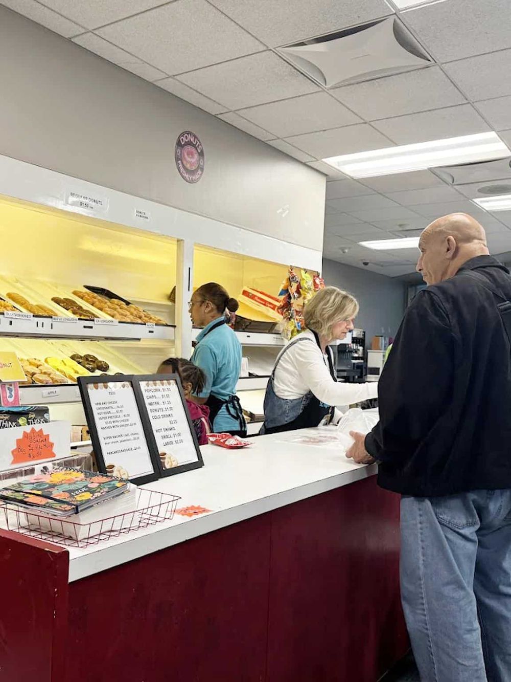 Freshly baked donuts at a bakery counter in a convenience store, with customers ordering and staff serving.
