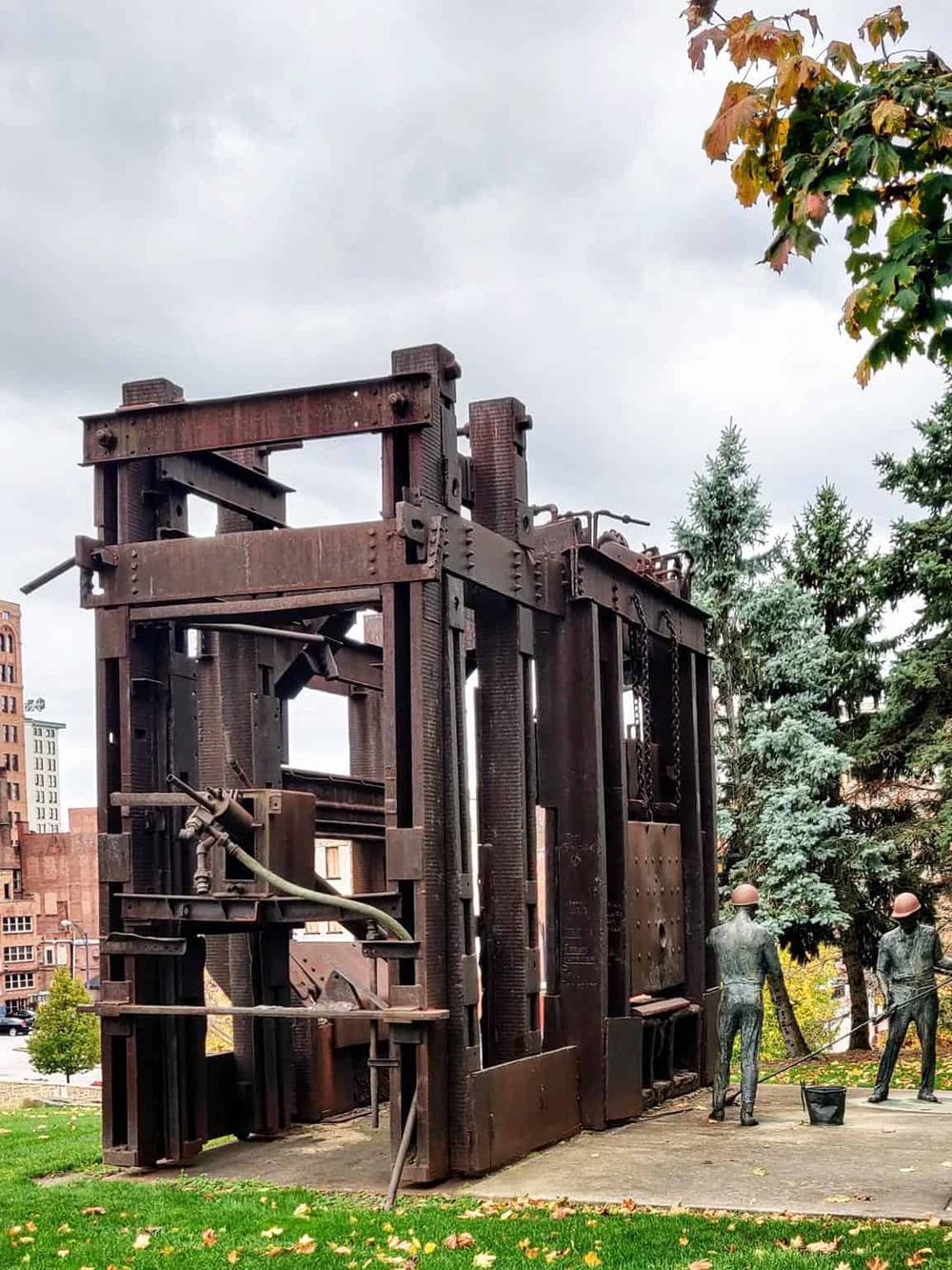Industrial sculpture outdoor art installation inspired by machinery, in a park with trees and cloudy sky.