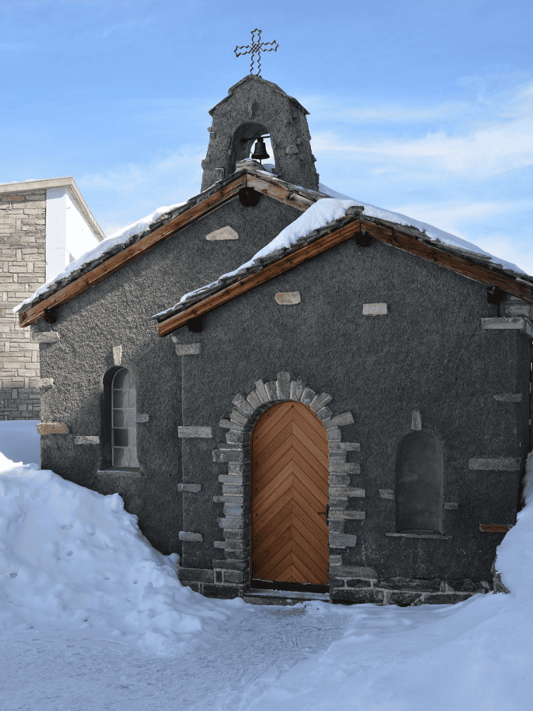 Small stone chapel with solar cross, snow, and wooden door in winter setting.