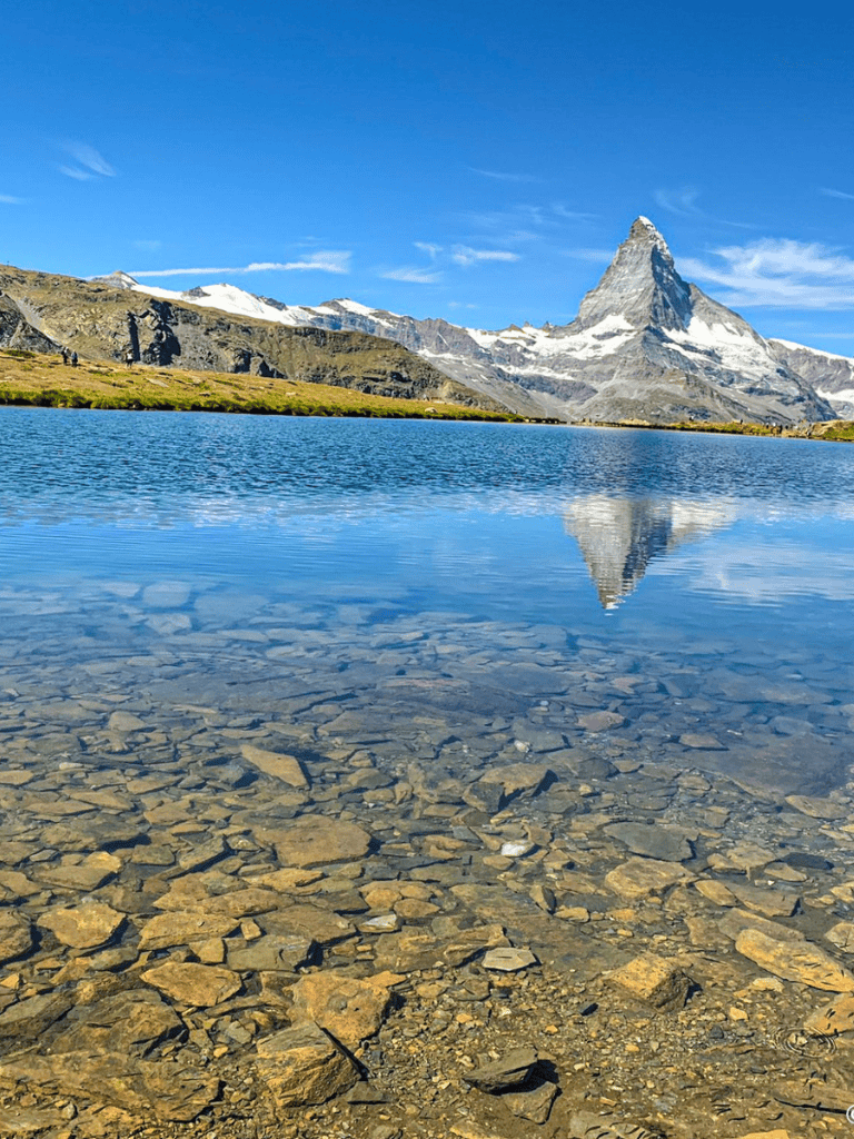 Serene mountain lake with reflection of snow-capped peaks, including the Matterhorn, under clear blue sky.