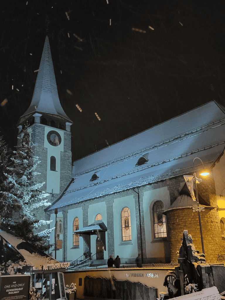 Snowy church with tall steeple and warm lit windows at night, under snowstorm, in a winter landscape.