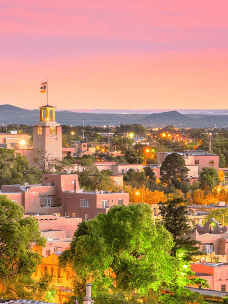 Colorful sunset over Santa Fe, New Mexico with adobe-style buildings and lush greenery.