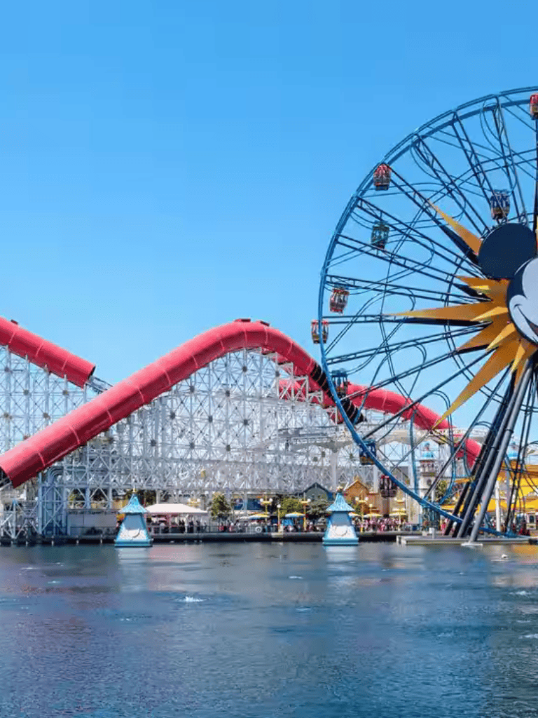 Colorful Ferris wheel and roller coaster at an amusement park near water under a bright blue sky.